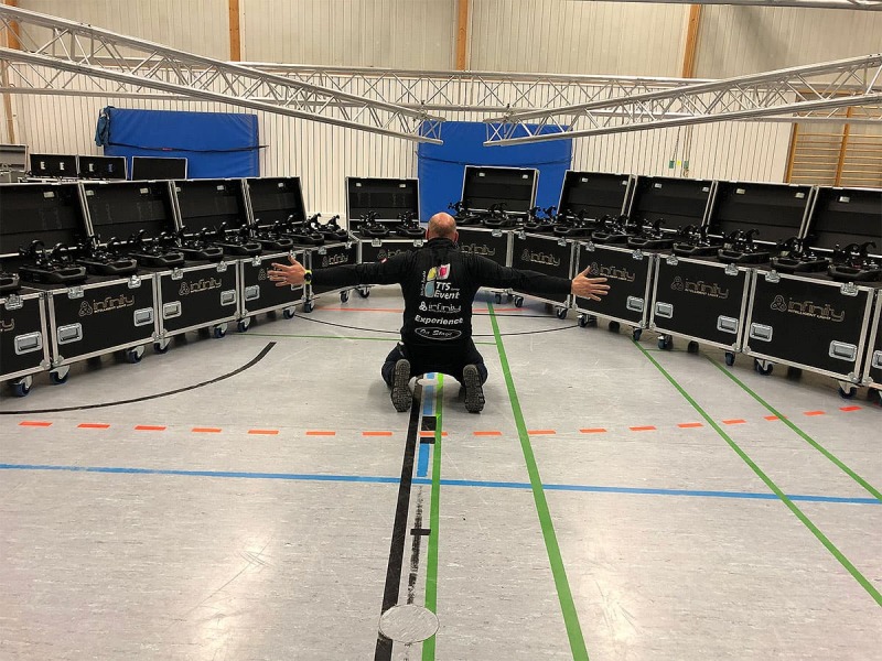 Mann kniet mit ausgestreckten Armen in einer Sporthalle vor einer Reihe halbrund angeordneter, offenen Rollkoffern mit Equipment darin