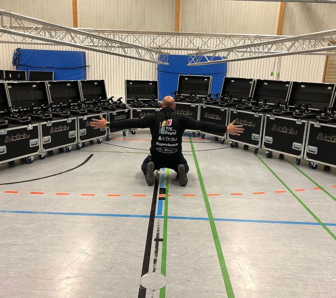 Mann kniet mit ausgestreckten Armen in einer Sporthalle vor einer Reihe halbrund angeordneter, offenen Rollkoffern mit Equipment darin