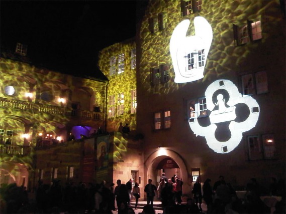 Ein Lichtspiel, bestehend aus verschiedenen Mustern und Symbolen, werden bei Nacht an eine Fassade projiziert